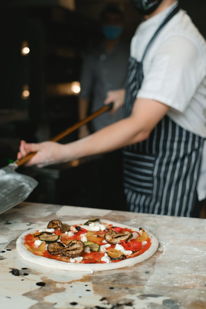 Chef in apron prepares an eggplant pizza inside a restaurant kitchen.